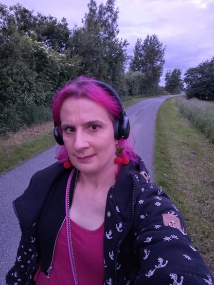Eine Frau mit pinken Haaren, Kirschohrringen, Kopfhörern auf einem schmalen Weg. Sie trägt pinkes Kleid und darüber einen schwarzen Bolero und eine weite, schwarze Jacke mit weißen Ankern. Auf beiden Seiten des Weges sind Wiese und Bäume. es dämmert