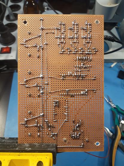A clamp with a piece of perfboard, showing the solder side of what seems to be an electronic circuit. Many solder points can be seen and some connections between them, made with tinned wire.