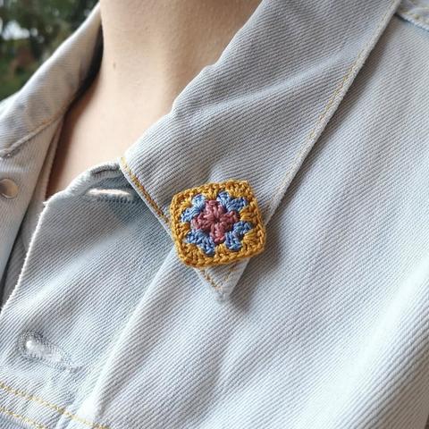Eine gehäkelte Brosche in Form eines Granny Squares am Kragen einer Jeansjacke.