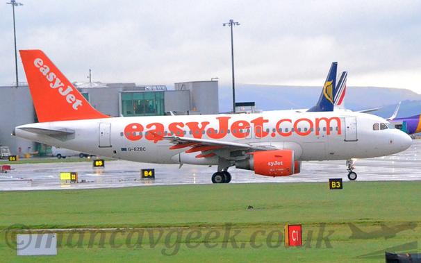 Side view of a twin engined jet airliner taxiing from left to right.
The plane is mostly white, with large , billboard-style orange "EasyJet[dot]com" titles on the fuselage, text repeated diagonally in white on the bright orange tail, and on the sides of the engine pods mounted under the wings, although the first 2 letters are missing on the engine.
Lush green grass fills the foreground, with a terminal building in the background on the left, and the tails of some other planes on the right.
Tree covered hills in the distance meet bright but hazy skies and cloud in the distance.