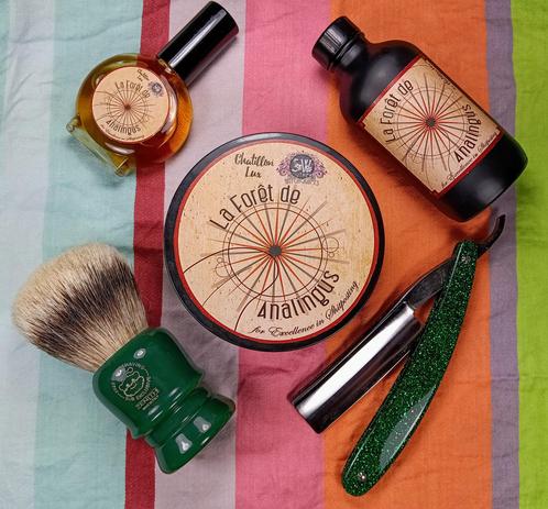 (Le Chat Mistral description)

The image showcases a collection of grooming items arranged on a striped cloth. At the centre, there's a round tin labelled "La Forêt de Analingus" with a decorative design. To the top left, there's a small bottle of cologne, and to the top right, a larger bottle of aftershave. On the bottom left, there's a green shaving brush with a badger hair brush head. On the bottom right, there's a green straight razor with a wooden handle. The items are neatly arranged, suggesting a traditional shaving setup.