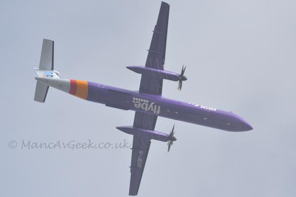 View from pretty much underneath of a high-winged, twin propellor-engined airliner flying from left to right at a low altitude, with flaps extended from the rear of the wings and undercarriage already retracted, suggesting it has just taken off.
The plane is mostly extremely purple, with a wide yellow and slightly narrower red band around the rear fuselage, and a white tail and tail-cone.
There are white "Flybe" titles over 6 white dots on the forward fuselage, repeated under the belly, and white text "Faster than road or rail" on the sides of the engines under the wings.
The white registration "G-JECF" is on the bottom of the left wing.
There are blue "Flybe" Titles on the tail.
Spinning vapour trails are visible around the engine pods, created by the propellor blades spinning through damp air.
Grey sky fills the rest of the frame.