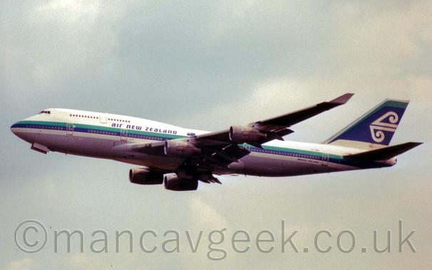 Side view of a very large, 4 engined jet airliner flying from right to left at a low altitude, with flaps extended from the rear of the wings, the undercarriage already tucked away, and the nose raised sharply, suggesting it has just taken off.
The plane is mostly white, with a grey belly, and a green and blue stripe running along the body, covering the lower deck cabin windows., with blue "Air New Zealand" titles at the rear of the upper deck.
The tail is mostly dark blue, with a green streak at the front and top, and a stylised white wave in the middle.
Grey cloudy sky with patches of blue fill the rest of the frame.