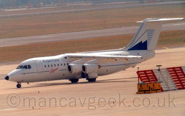 Side view of a high-winged, 4 engined jet airliner taxiing from right to left, with flaps extended from the rear of the wings, suggesting it is going out to a runway for takeoff.
The plane is mostly white, with a thin blue stripe running along the body, overlaid with diagonal red "Alpine" titles below the passenger cabin windows just ahead of the wings, and blue "Flightlines" titles on the upper forward fuselage.
The black registration "G-HWPB" is on the upper rear fuselage, partially obscured by the wing.
The tail is white, with a blue and white stylised image of a snow-capped mountain, with short, light blue lines on it's leeward side.
Grey concrete apron fills the foreground, with an angled red and white metal barrier on the right of the frame, while wide strips of green grass line a black runway in the background.