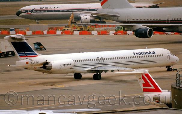 Side view of a twin engined jet airliner with the engines mounted on the sides of the rear fuselage, facing to the right but being pushed back to the left by an out-of-frame tug attached to the planes nosewheel.
The plane is mostly white, with a trio of dark blue pinstripes running along the lower fuselage under the passenger cabin windows, and dark blue "Centennial" titles on the upper forward fuselage, repeated on the sides of the engine pods.
The black registration "EC-FVV" is on the upper rear fuselage, next to a small image of a Spanish flag, just aft of the engines.
The tail is mostly dark blue, with 2 thin lighter blue stripes and a similar red stripe at the bottom, all outlined in white.
The foreground is mostly filled with grey concrete apron, with the white and red tail of a small jet airliner poking out from behind  the corner of a dark grey terminal engine on the bottom right of the frame.
A grey and dark blue, very large 4 engined airliner is taxiing across the frame from left to right in the background, with a white and black 3 engined jet airliner taxiing from right to left towards the runway beyond that.