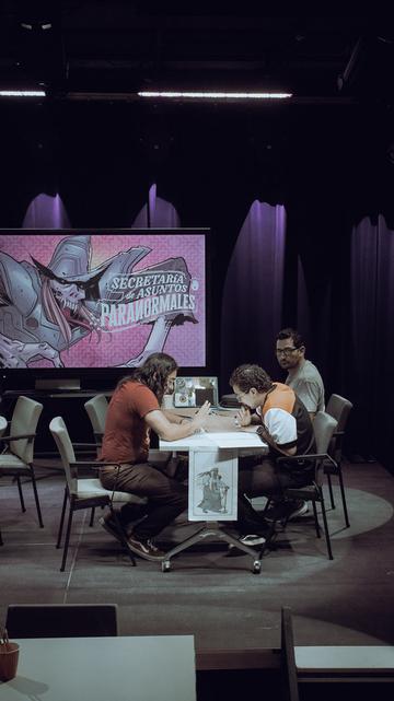 Tres personas sentadas alrededor de una mesa jugando una partida de rol. Al fondo hay una pantalla con una ilustración del juego "Secretaría de Asuntos Paranormales" desarrollado por la comunidad Leyendas del Noreste del Laboratorio Cultural Ciudadano de Monterrey (México).