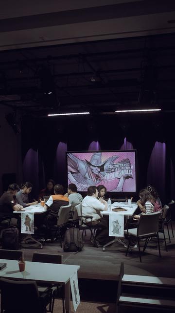 Dos mesas con varias personas sentadas alrededor mientras juegan una partida de rol. Al fondo hay una pantalla con una ilustración del juego "Secretaría de Asuntos Paranormales" desarrollado por la comunidad Leyendas del Noreste del Laboratorio Cultural Ciudadano de Monterrey (México).