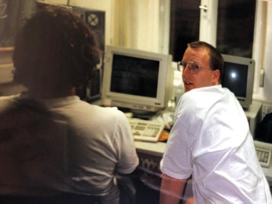 Two guys sitting in front of a CRT monitor, one of them turning his head to the camera.