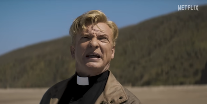 Image from the Forbes article, showing Rhys Darby in “Love, Death + Robots”. He is seen from the shoulders up, standing on a beach and squinting up at something above the camera with a puzzled or shocked look on his face. He wears a black shirt with a clerical collar, and a tan jacket.