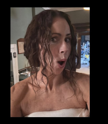 Selfie of Minnie Driver looking at the camera with her face turned to the side, her mouth open and eyes wide as if in surprise. Her hair looks damp, with some strands falling into her face while the back of her hair is tied up. Her shoulders are bare and she has a towel or sheet wrapped around her.