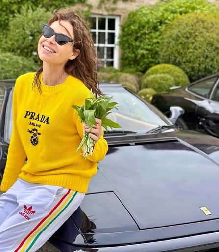 Minnie Driver smiles broadly with her head thrown back while leaning on the hood of a black Ferrari car and holding up a bunch of wild garlic like a bouquet. She wears dark cat-eye sunglasses, a yellow Prada sweater and white Adidas sweatpants.