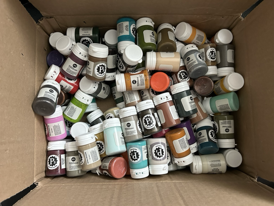Photo of an open box full of different paint pots with flip-caps. They’re all tossed around haphazardly.