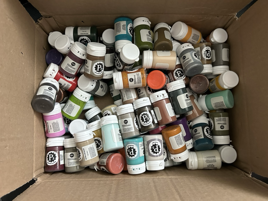 Photo of an open box full of different paint pots with flip-caps. They’re all tossed around haphazardly.