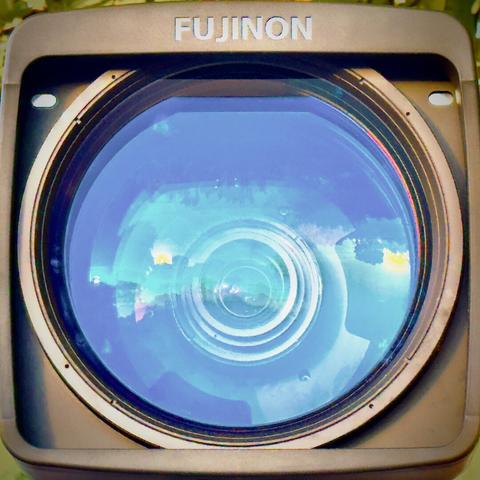 The front element of a large broadcast lens reflects the morning sunlight, the word "FUJINON" etched into the top of its hood.