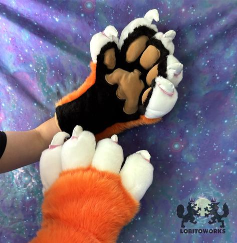 Orange, white, and black fursuit hands with big padded fingers and large white claws with pink cuticles. The pawpads are tan in color and stuffed so they stick out, and the palms are black. The hands are displayed in front of a blue-violet space themed backdrop.