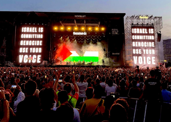 Photo at a large outdoor concert at dusk. Image shows the back of thousands of people, many holding up their phones, as they look towards the stage where a hume Palestinian flag is lit up. Lights above spell out Fontaines DC. Large lit up signs on both sides of the stage spell out: ISRAEL IS COMMITTING GENOCIDE USE YOUR VOICE