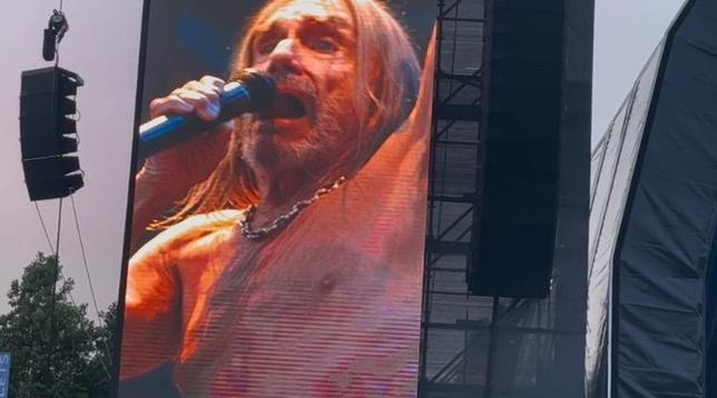 This image shows a close-up view of a large video screen at an outdoor concert venue. The screen displays a vivid, high-resolution image of a performer singing into a microphone. The person on screen has long, light-colored hair and appears to be shirtless, captured mid-performance with an intense, energetic expression typical of rock concerts.

The video screen itself is quite large and appears to be part of the stage setup, with visible metal framework and rigging structure around it. To the left side of the frame, you can see part of the concert’s sound equipment, including what looks like a speaker array or lighting rig.

The setting appears to be daytime at an outdoor festival or concert venue, with some trees visible in the background and natural lighting. The screen’s bright display contrasts against the daylight sky, allowing the audience to clearly see the performer’s close-up image even from a distance.

This type of large video screen is common at major concerts and festivals, ensuring that audience members far from the stage can still see detailed views of the performance.​​​​​​​​​​​​​​​​