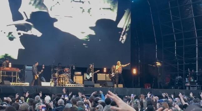 This image shows a live concert performance taking place on an outdoor stage. The stage features a band with several musicians - there’s a drummer at a kit in the center-back, guitarists positioned on the left and right sides of the stage, and what appears to be the lead performer (Iggy Pop) on the right side of the stage. The stage setup includes various amplifiers and musical equipment typical of a rock concert.

Above the performers, there’s a large video screen displaying what looks like projected visuals or possibly live footage of the performance. The stage is covered by a large tent or canopy structure with visible support beams creating geometric patterns against the sky.

In the foreground, there’s a enthusiastic crowd of concert-goers with many people raising their hands and phones in the air, clearly engaged with the performance. The audience fills the visible area in front of the stage, creating the typical atmosphere of a lively outdoor music festival or concert venue.

The lighting suggests this is taking place during daytime or early evening, with stage lights providing illumination for the performers against the natural outdoor setting.​​​​​​​​​​​​​​​​