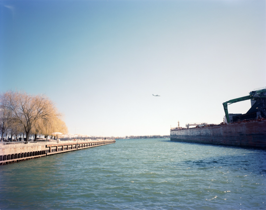 The harbour between two quays: the one on the left has been converted to a park with beach and pink umbrellas; on the right a cargo ship is being unloaded. A plane flies low for a landing.