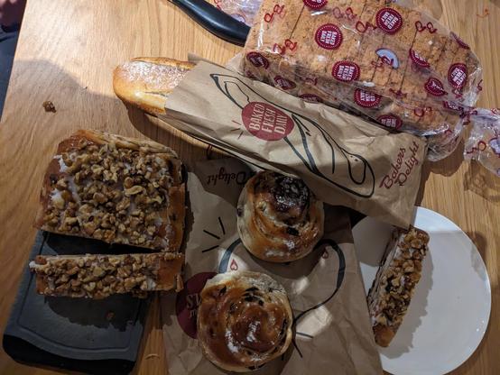 a Walnut log on a chopping board, with a slice on a plate.  There are two apple scrolls on a brown paper bag, a loaf of bread in a plastic bag and a long baton in a paper bag.