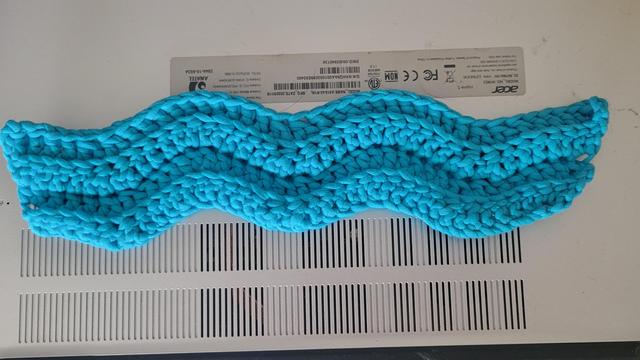 A10 inch by 3 inch bolt of cloth lying on the back of a silver laptop. The cloth is crocheted using a sky blue cotton yarn, and has a wavy Chevron pattern to it.