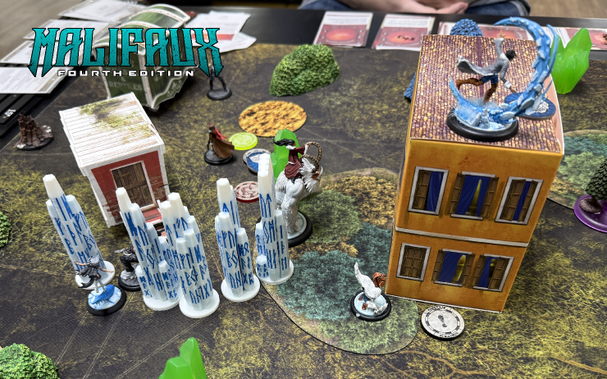 Picture of a Malifaux game in progress, showing a wall of ice pillars blocking a path between two terrain buildings with friendly minis in the foreground and enemy minis behind the wall.
