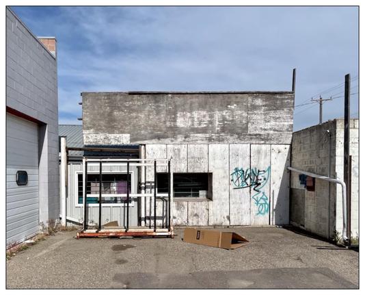 Weathered building with a flat roof, featuring a patched-up facade of white and gray panels. The front of the building includes a set of metal-framed glass doors beneath a faded sign, barely visible. To the right, graffiti in blue and green adorns the wall. A fallen cardboard box lies on the concrete foreground, and adjacent structures partially enclose the scene under a clear sky.