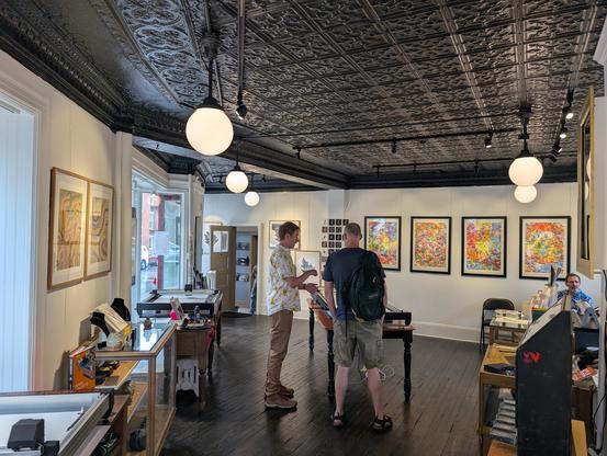 Paul speaking to a visitor in a gallery featuring large colorful pen plotter drawings on the walls
