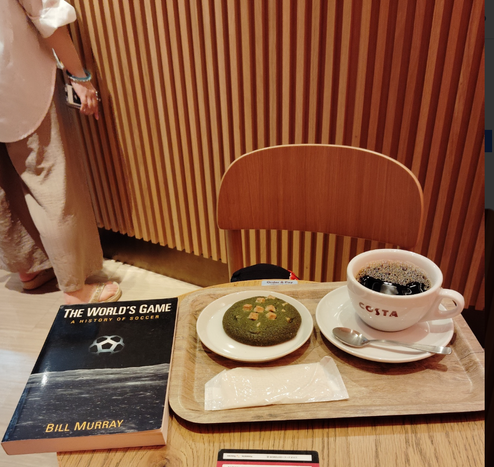 The black paperback with a cover of a soccer ball as earth over the moon is on the left edge of the round wooden table. A tray is to the right with a green cookie with white chocolate chips on a white plate in front of a napkin and a white mug and saucer and silver spoon of black coffee and COSTA in red on the mug. The legs of a patron can be seen by the corrugated wooden counter