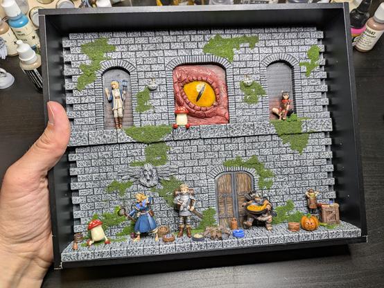 The dungeon backdrop in a small black shadowbox insert, held by a hand. 4 main characters and a monster are on the ground floor, with cooking implements scattered around. On the top floor are 2 additional characters, plus a walking mushroom that's sitting down in front of a gigantic red dragon's eye, poking through an open arch.