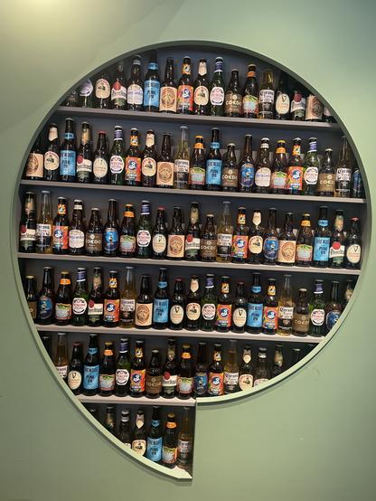 Green painted wall with a round quote mark cut out to recessed shelving filled with colourful beer bottles - about ten different brands including Japanese, Belgian, America, with my eye drawn to the pale blue of the UK’s Brew Dog Punk IPA.