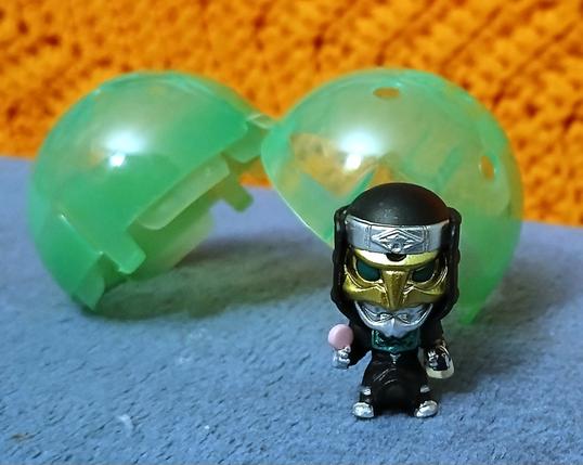 A mini figure of Deneb from Kamen Rider Den-O propped up by an open gashapon capsule