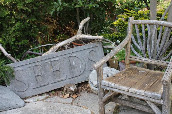 A rustic wooden chair on a flagstone patio with a cement sign SEEDS next to it with tree branches behind it  and a large stone and  cement bowl with cement item set in it. There are trees and plants in the background.