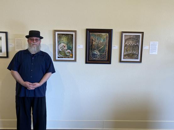 Photograph of a bearded man in a pork pie hat standing next to three artworks on a gallery wall.