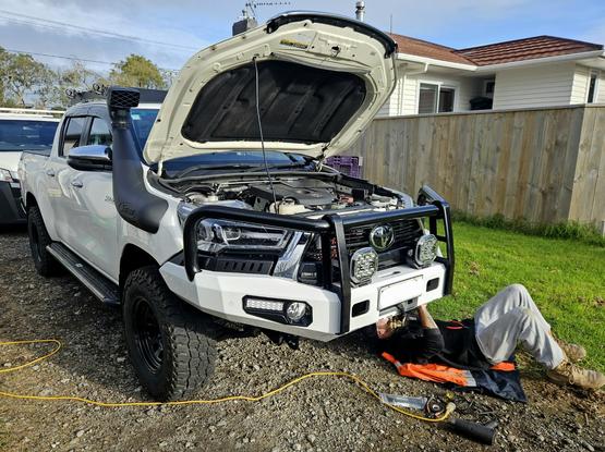 White Toyota Hilux double cab (4 doors with shorter tray) with black steel wheels and mud terrain tyres, on a gravel driveway in suburbia. The neighbours lawn and house visible right behind, with my brother laying under the front of the vehicle cable tying up the wiring to the park and indicator lights. There are also large spotlights mounted in front of the grill.