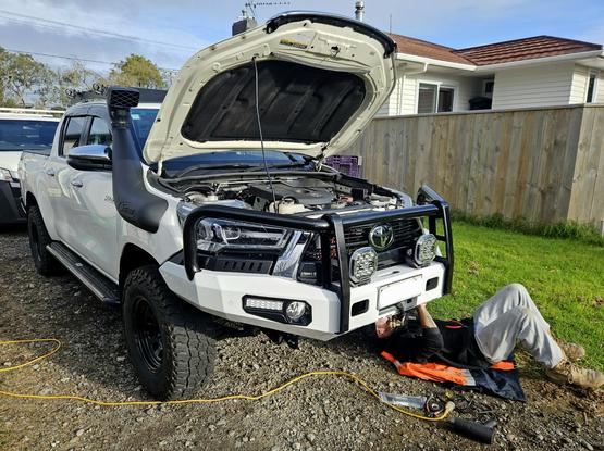 White Toyota Hilux double cab (4 doors with shorter tray) with black steel wheels and mud terrain tyres, on a gravel driveway in suburbia. The neighbours lawn and house visible right behind, with my brother laying under the front of the vehicle cable tying up the wiring to the park and indicator lights. There are also large spotlights mounted in front of the grill.