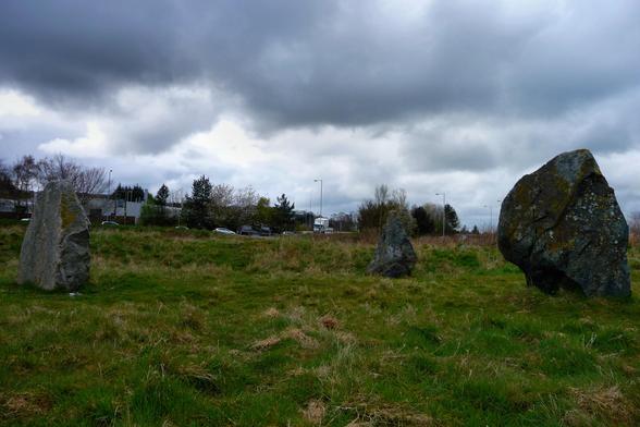 Three large standing stones inside a grass-covered circular earthen bank. The stone on left is carved with Pictish symbols and is not original to the monument. It's very overcast with heavy grey rainclouds filling the sky.