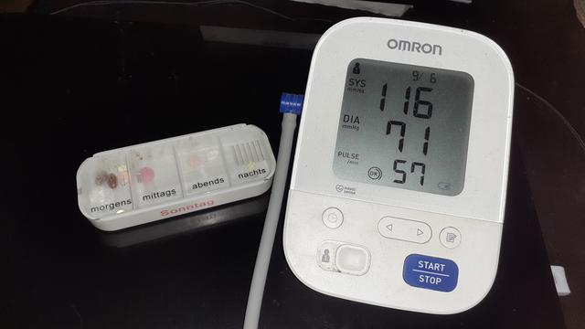 Rechts im Bild steht ein Oberarmmanschetten Blutdruckmessgerät und links daneben liegt eine Tages-Pillendose. Beides steht/liegt auf einem dunkelbraunen Couchtisch und beide Dinge sind weiß.