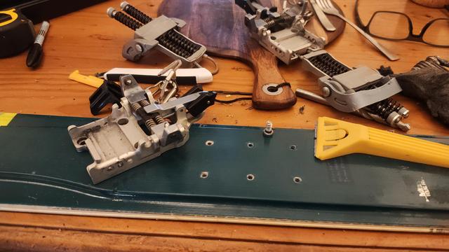 A cluttered table containing parts of telemark ski bindings, scraper blades, a drill bit, and part of a ski with holes drilled in it