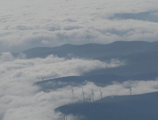 This photo captures a breathtaking aerial view of a mountain range partially covered by a thick blanket of clouds or fog. Peeking through the mist are several wind turbines scattered across the ridges, their blades still or gently turning. The scene has a serene, almost surreal quality, with the dark blue-green tones of the mountains contrasting against the soft, white cloud cover. The wind turbines add a subtle element of human presence and renewable energy infrastructure amidst the natural landscape.