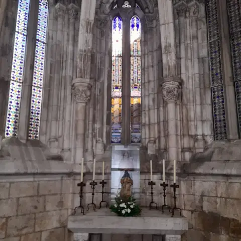 A long stained glass window at the end of a nook in a church. The walls are grey stone and there is a table in front of the window with the Virgin Mary, some flowers and 6 candles surrounding her. The main stained glass starts yellow at the bottom and moves to blue at the top. It depicts some columns. The two to each side are just matching patterns.