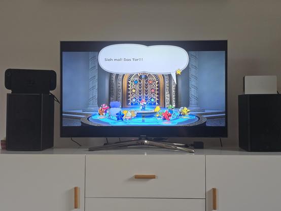 Das Bild zeigt einen Fernseher auf einem weißen Fernsehschrank. Links und rechts vom Fernseher stehen schwarze Audio-Boxen. Auf den Boxen stehen links eine schwarze Switch 2 im Dock und auf der rechten Seite nur ein weißes Switch-1-Dock.
Auf dem Bildschrim ist eine Szene aus dem Spiel Paper Mario: Die Legende vom Äonentor zu sehen.