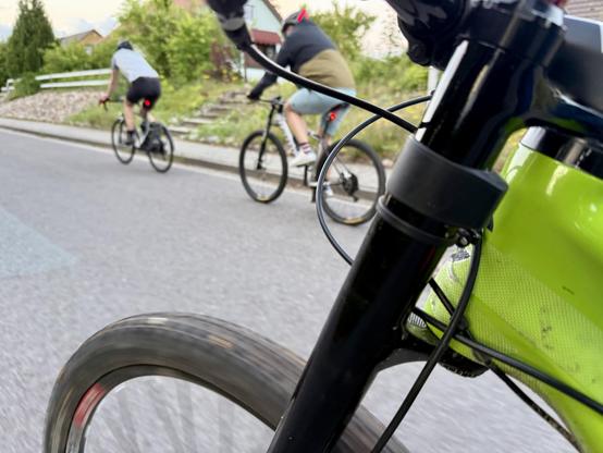 Eine Nahaufnahme der lefty Vordergabel und des Rades eines Fahrrads auf einer Fahrbahn. Im Hintergrund sind zwei Radfahrer zu sehen, die in die gleiche Richtung fahren.