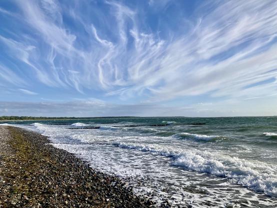 In der unteren Fotohälfte graublaues Meer mit Wellen, Gischt und einem breiten Streifen Spülsaum an steinigem Strand. Darüber ein hellblauer Himmel mit vom Wind zerzausten Wolkenstreifen, die strahlenförmig vom Bildmittelpunkt nach oben verlaufen.