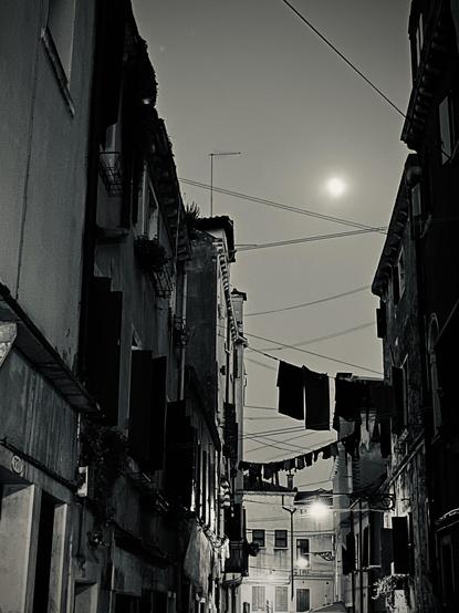 Black and white photo taken at night, looking slightly up and along a narrow Venetian street, criss-crossed by a tangle of washing lines at various angles, some with laundry on them and some without, which look to me like the complex rigging of a mad imaginary boat with odd towel- and trouser-shaped sails. The houses on either side of the street are in shadowy darkness, you can just make out the shapes of windows and shutters, but more as texture rather than detail. There are also some power lines strung across the street, and there are three bright lit streetlights in a gentle curve shape rising from the centre bottom of the frame, to slightly right-of-centre about a third of the way up the frame; the eye is drawn upwards to the fourth point of light, the almost-full moon, a third of the way from the top of the frame. The image feels mysterious and secretive.
