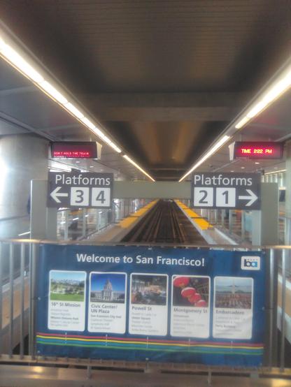A sign indicates platforms 2 and 1 to the right. Another sign indicates platforms 3 and 4 to the left. There is a track leading away from us with yellow safety strips on both sides. There is a promotional sign "Welcome to San Francisco" with sections on 16th St Mission, Civic Center/UN Plaza, Powell Street, Montgomery Street, and Embarcadero. An overhead sign to the right of the track reads "Time 2:22 PM." An overhead sign to the left of the trark reads "Don't hold the train."