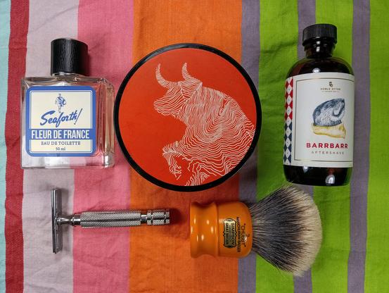 (Le Chat Mistral description)

The image showcases a collection of grooming items arranged on a striped background. At the top left, there's a bottle of Seaforth! Fleur de France Eau de Toilette, which is a light blue and white bottle. In the centre, there's a round container with a red lid featuring a white topographic map design. To the right, there's a brown bottle of Noble Otter Barrbarr Aftershave with a label depicting a beaver on a piece of wood. Below these items, there's a silver safety razor on the left and a badger hair shaving brush with an orange handle on the right. The colours and arrangement give a vibrant and organised feel.