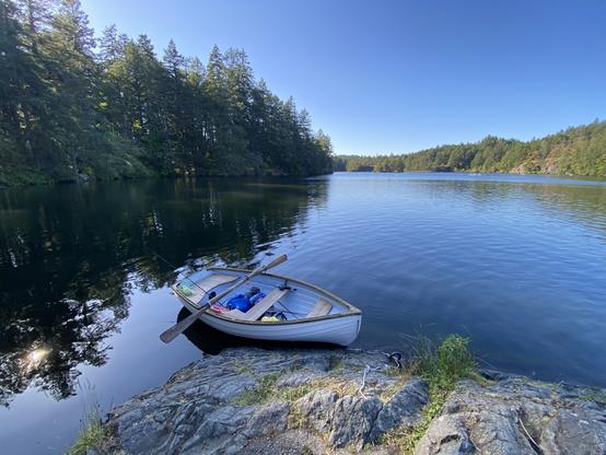 A white rowboat, 9' long, in the style of a Whitehall Spirit Minto 9, is floating beside the shore at Thetis Lake. Trees and blue skies reflect off the lake.
