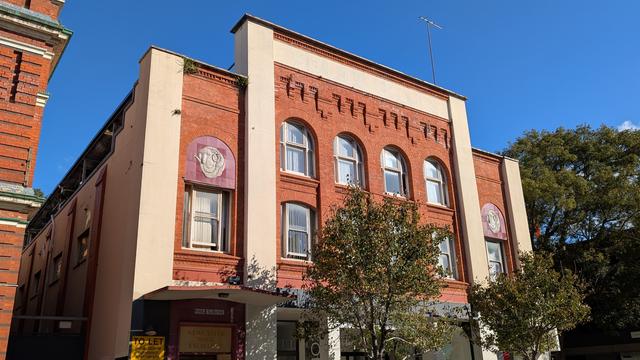 A historical red brick building in Newcastle, Australia. The Newcastle Wool Exchange. A square central frontage with 2 rows of 8 arched windows is framed at the top and side by flat cream concrete. Either side are shorter rectangles of red brick frontage, with a single arched window and the merino motif above, and thick vertical borders of the same flat cream concrete.