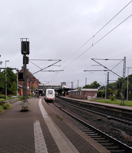 You are looking head on at the head of a Deutsche Bahn ICE train just arriving at a platform of a suburbian trainstation. Red brick station buildings, overhead wires and signals.